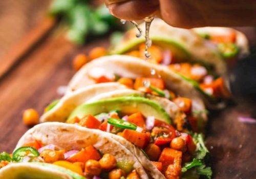 A hand squeezes lime over tacos filled with chickpeas, avocado, cilantro, and diced vegetables on a wooden board; lime slices nearby.