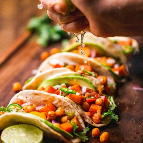 A hand squeezes lime over tacos filled with chickpeas, avocado, cilantro, and diced vegetables on a wooden board; lime slices nearby.