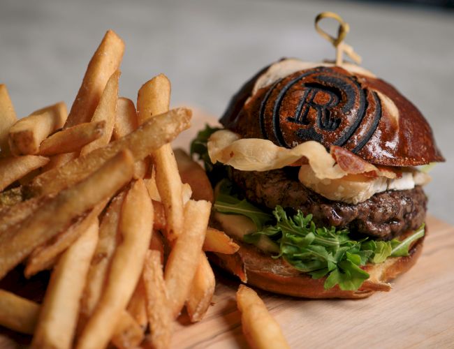 A burger with lettuce and cheese in a pretzel bun, alongside a serving of fries on a wooden board.