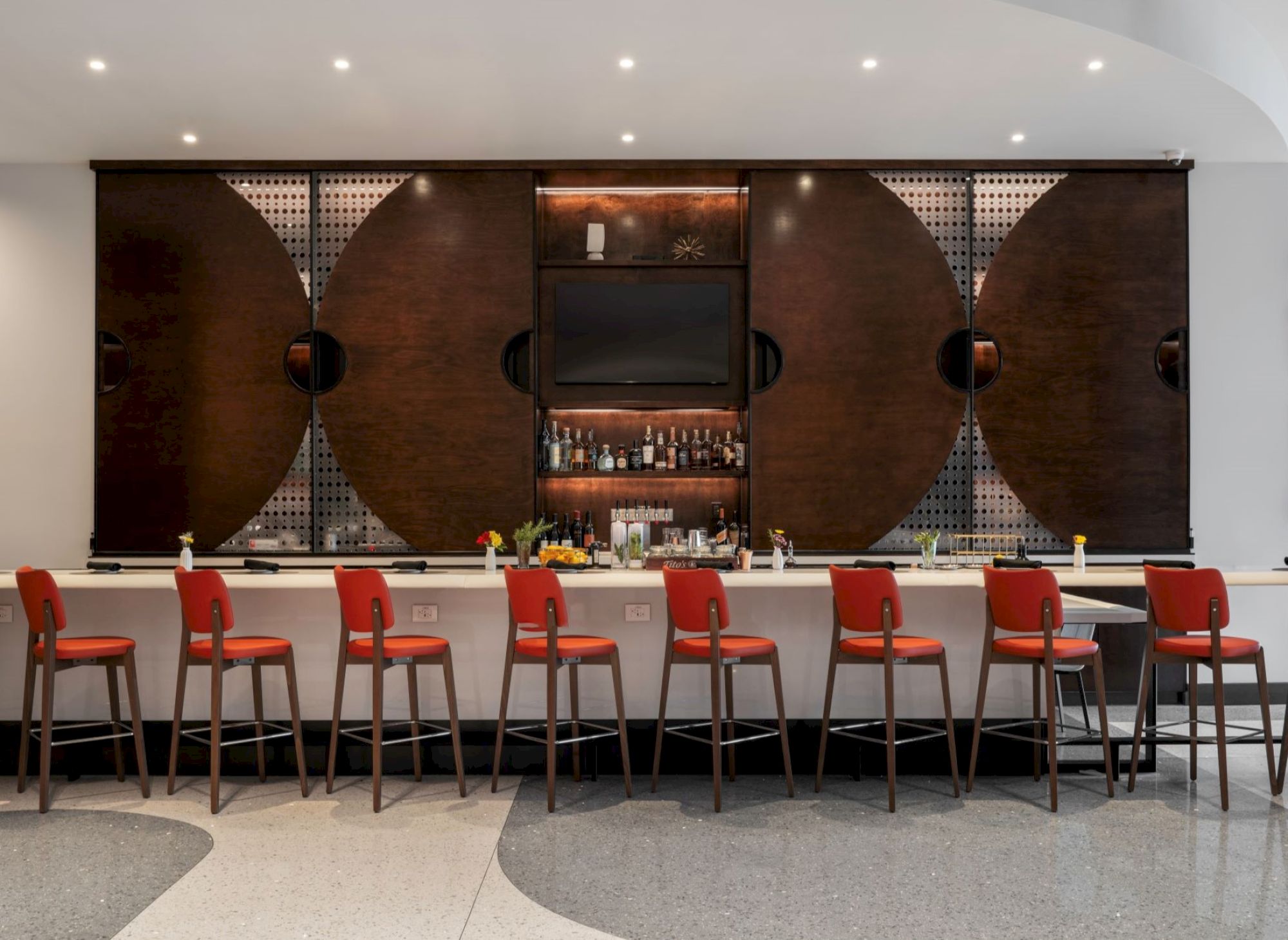 A modern bar with red stools and a stylish wooden backdrop, featuring a variety of bottles and a sleek design.