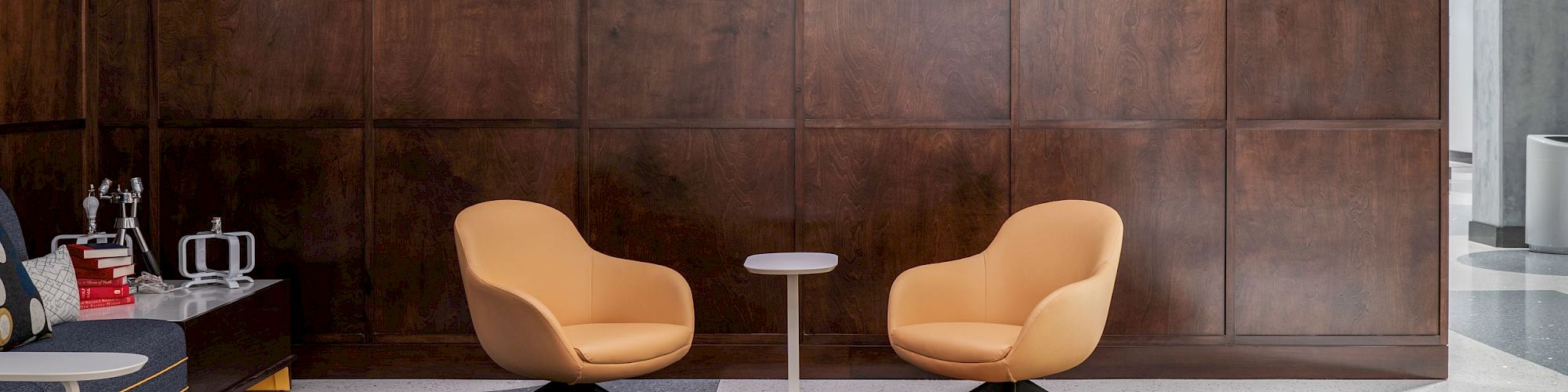 A modern lobby with wood paneling, two chairs, a small table, and a patterned rug. A sign reads "THE ROSENWALD COURT" on the wall.