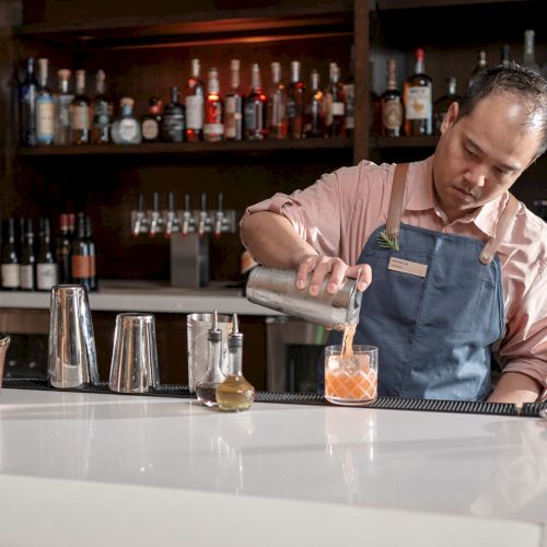 A bartender in a pink shirt and blue apron pours a drink into a glass at a bar with bottles and shakers in the background.