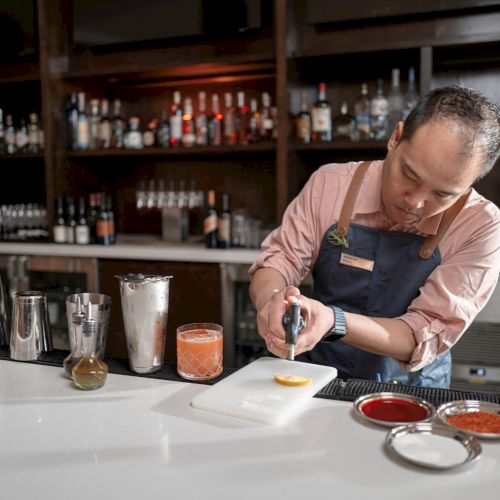 A bartender is preparing a drink, using a torch on a citrus slice at a bar with various bottles and tools on the counter.