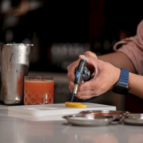 A person uses a torch to heat an orange slice at a bar. Nearby are a cocktail, shaker, and metal tools on a white surface.