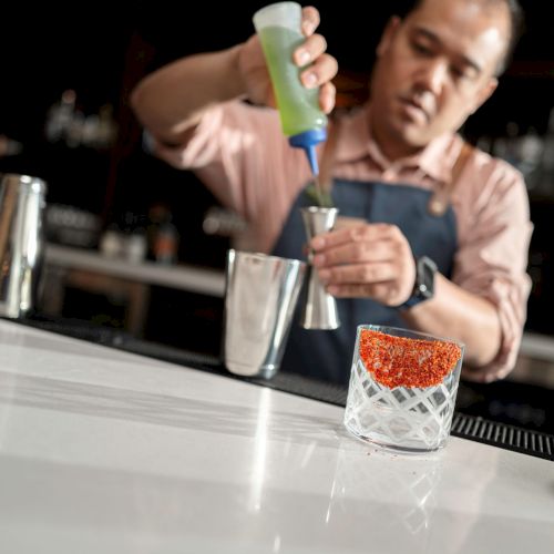 A bartender is preparing a drink, pouring liquid from a green bottle into a jigger, with a glass filled with red garnish on the bar counter.