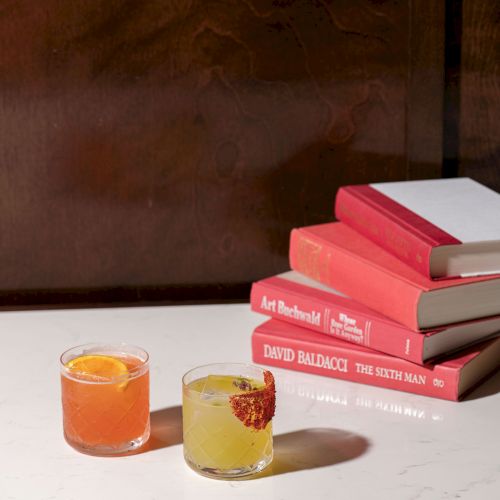 Two drinks on a table next to a stack of red and white books, set against a dark background.