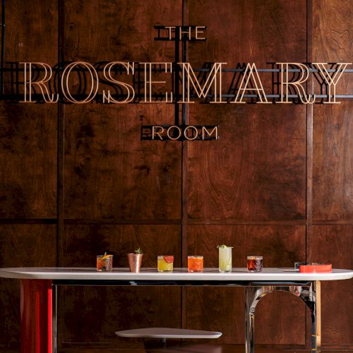 The image shows a table with various drinks in front of a wooden wall, with the sign "THE ROSEMARY ROOM" displayed above.