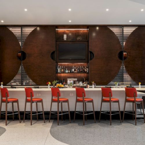 A sleek bar with nine red chairs, a modern design, and various bottles displayed on the counter and shelves behind it.