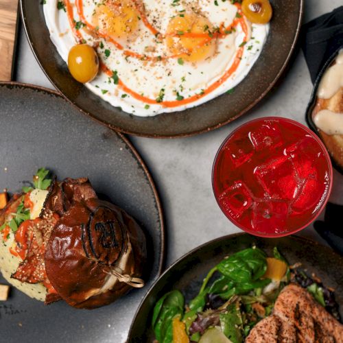 A spread of dishes including a burger, salad, roasted potatoes, a dessert, a creamy dish, and a red iced drink is shown.