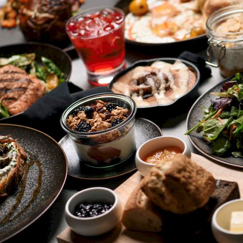 A variety of dishes including salads, bread, yogurt parfait, grilled items, and beverages are artfully arranged on a table.