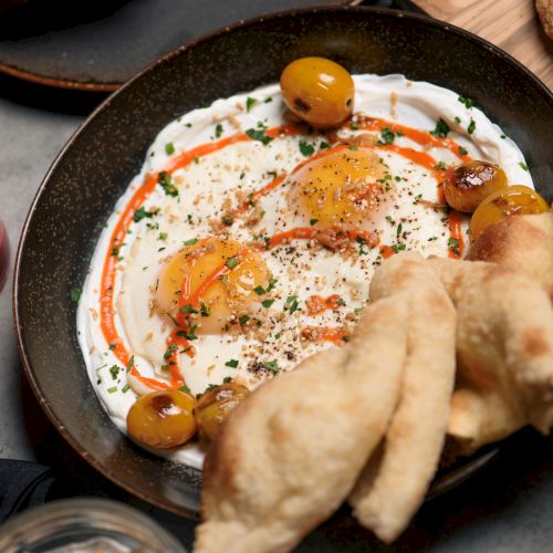 A dish with creamy sauce, eggs, olives, herbs, and bread on the side, accompanied by red and orange drinks.