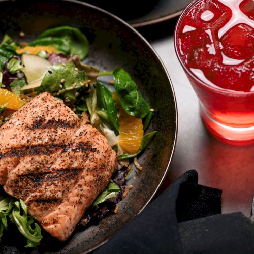 Grilled salmon on mixed greens with orange slices, a cold red beverage with ice, and a partial view of a dessert topped with sauce.