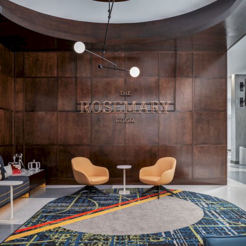 A modern lobby with wood paneling, two orange chairs, a round table, and a patterned carpet. A chandelier hangs from the ceiling.