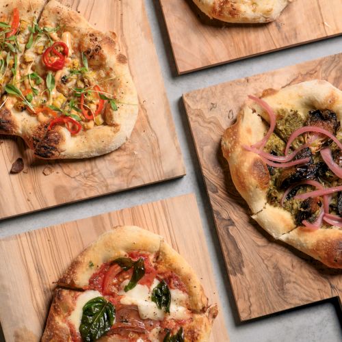 Various flatbreads with toppings like tomatoes, peppers, mushrooms, and onions on wooden boards.