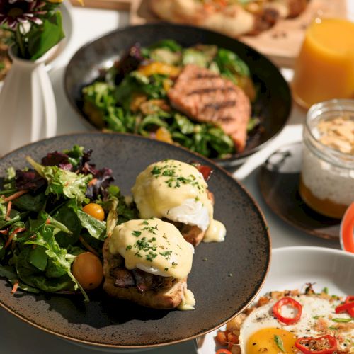 The image shows a variety of dishes, including salads, eggs benedict, grilled salmon, and a jar dessert with juice in the background.