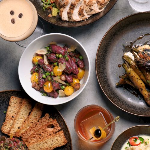 An assortment of dishes and drinks including salad, bread, roasted vegetables, cocktails, and a bean dish, displayed on a table.