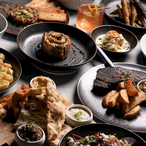 An assortment of dishes including pasta, bread, roasted veggies, and meat with sides, garnished and elegantly presented on a table.