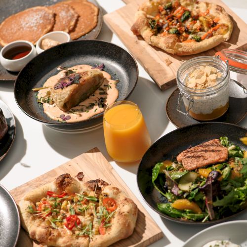 An assortment of delicious dishes including pizzas, salads, pancakes, and a glass of orange juice, arranged on a table with flowers.