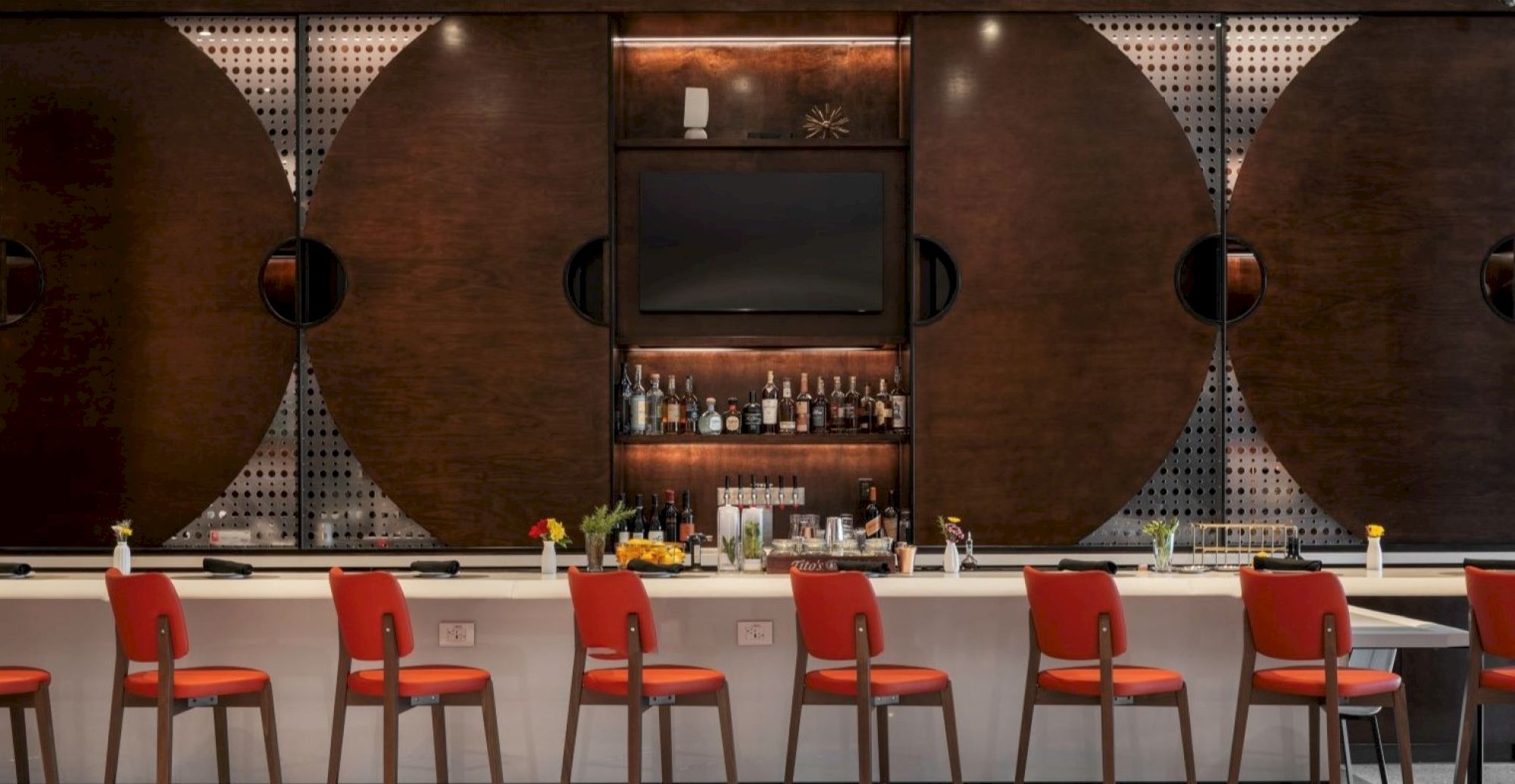 The image shows a stylish bar with red chairs, a wall-mounted TV, and a shelf stocked with bottles, set against a modern geometric backdrop.