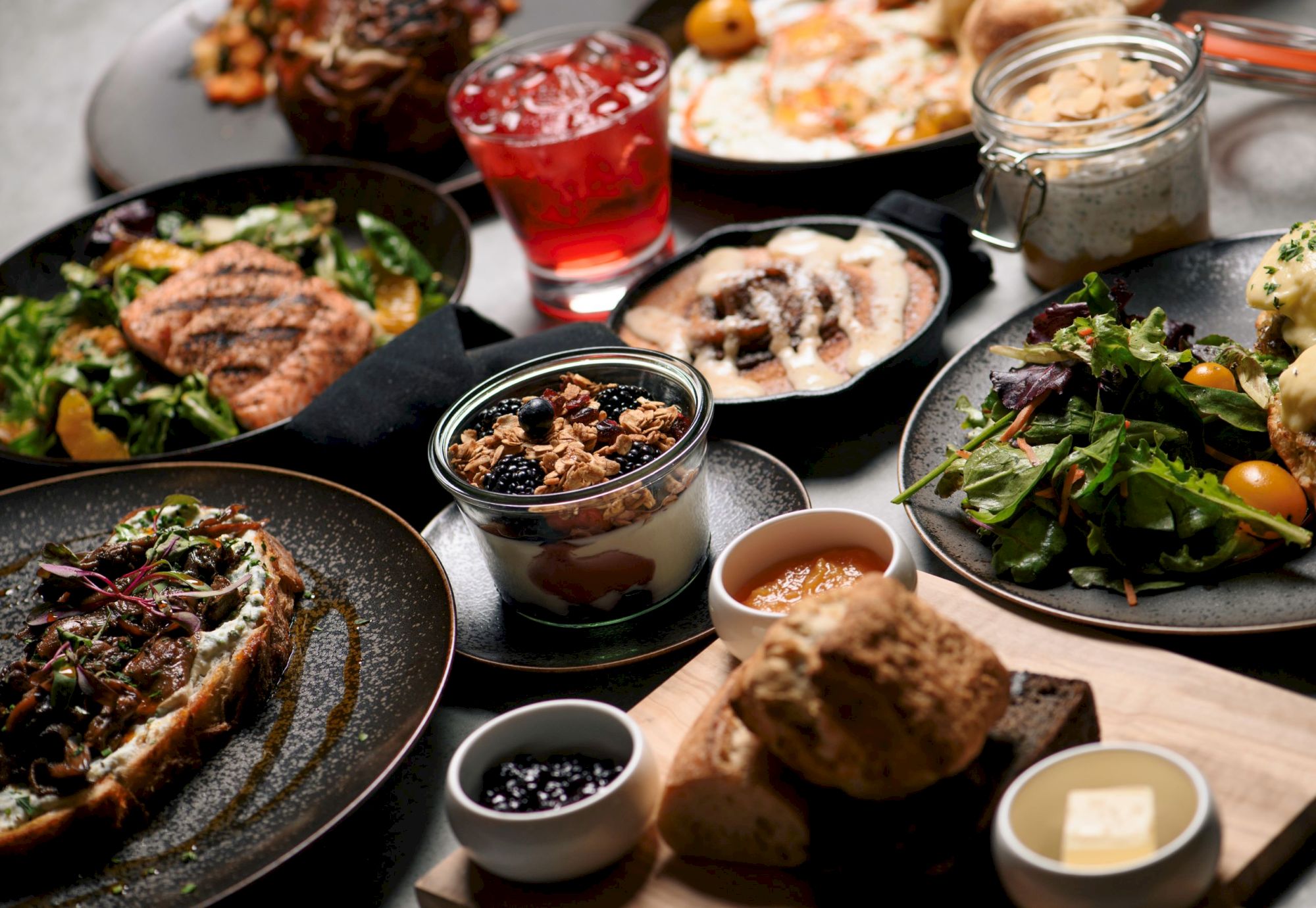 A spread of dishes including salads, toast, yogurt with granola, bread with butter and jam, and a cocktail.