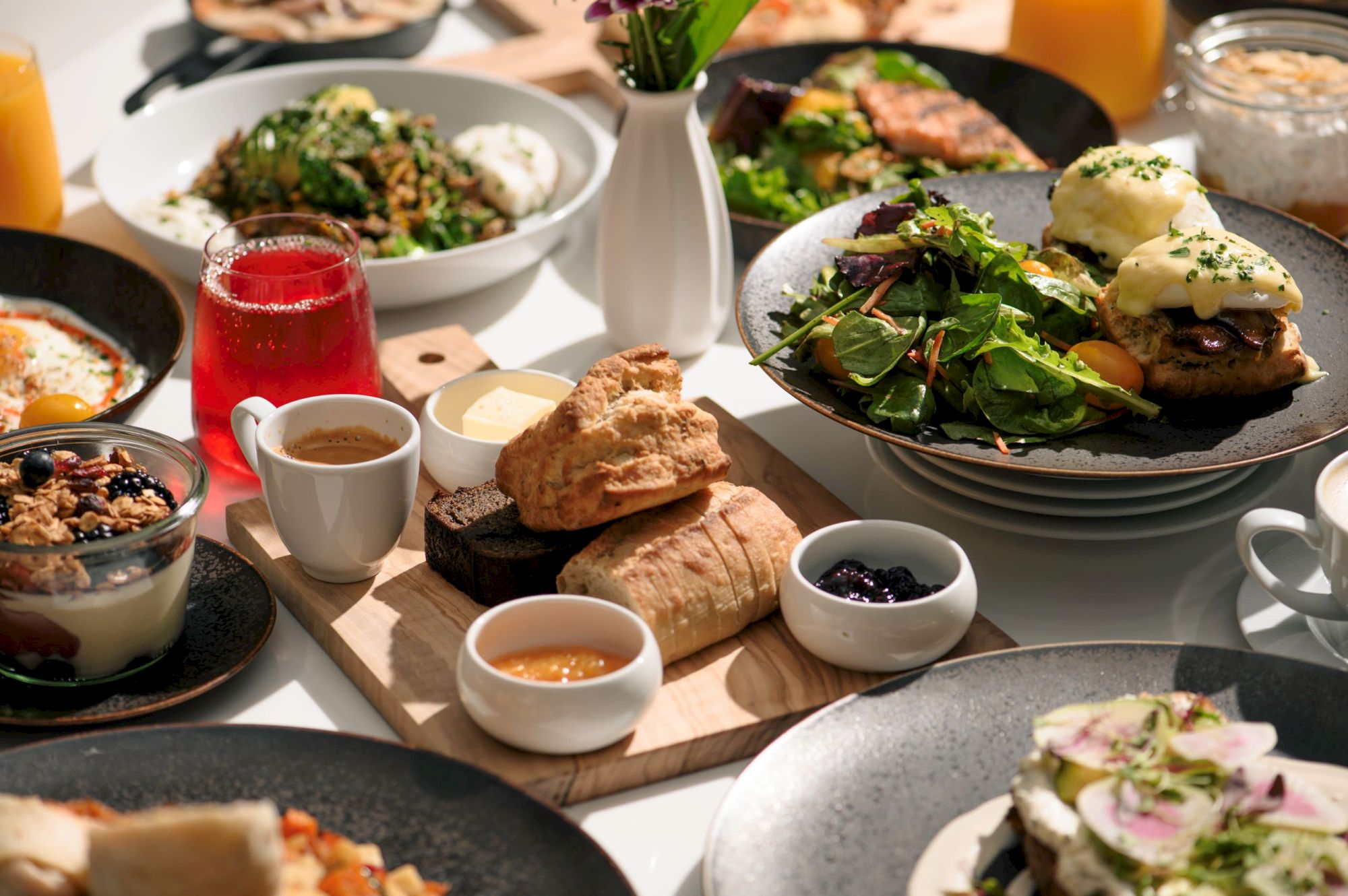 A variety of breakfast dishes including eggs Benedict, salad, pastries, granola, and drinks on a table.